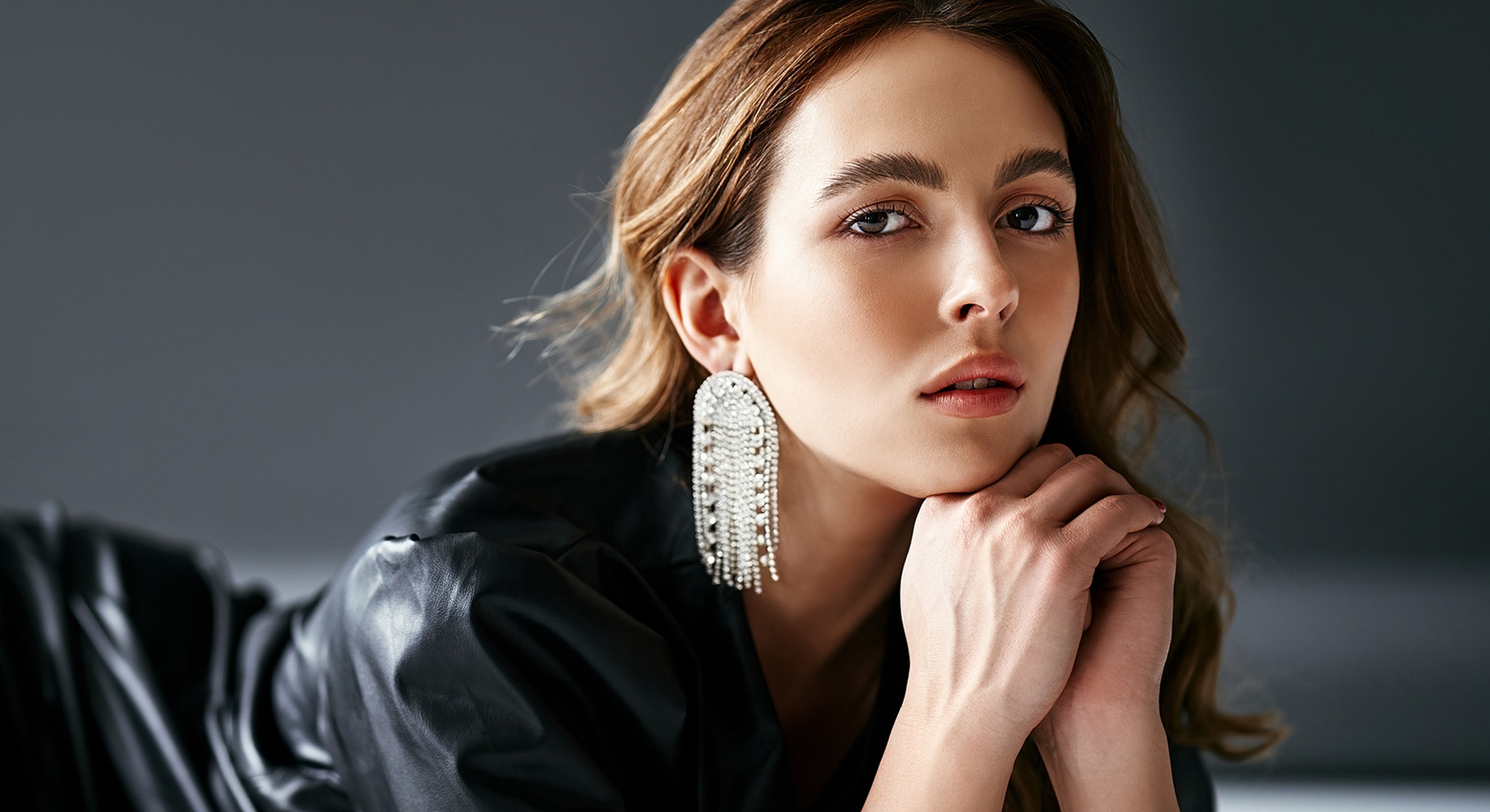 Woman with earrings posing thoughtfully against gray backdrop.